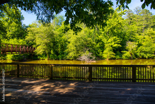 a brown wooden deck over the silky brown waters of Little River surrounded by lush green trees, grass and plants with a metal rust colored bridge and blue sky at Olde Rope Mill Park in Woodstock photo