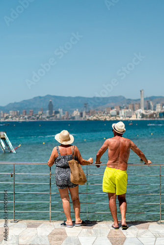 Rear view of a mature man and woman, wearing beachwear, looking at the sea and Benidorm beach from a metal railing.