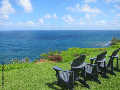 Relaxing concept. Three blue wooden chairs on a cliff s green lawn facing a deep blue ocean. Blue sky with white clouds. Princeville  Kauai  Hawaii.