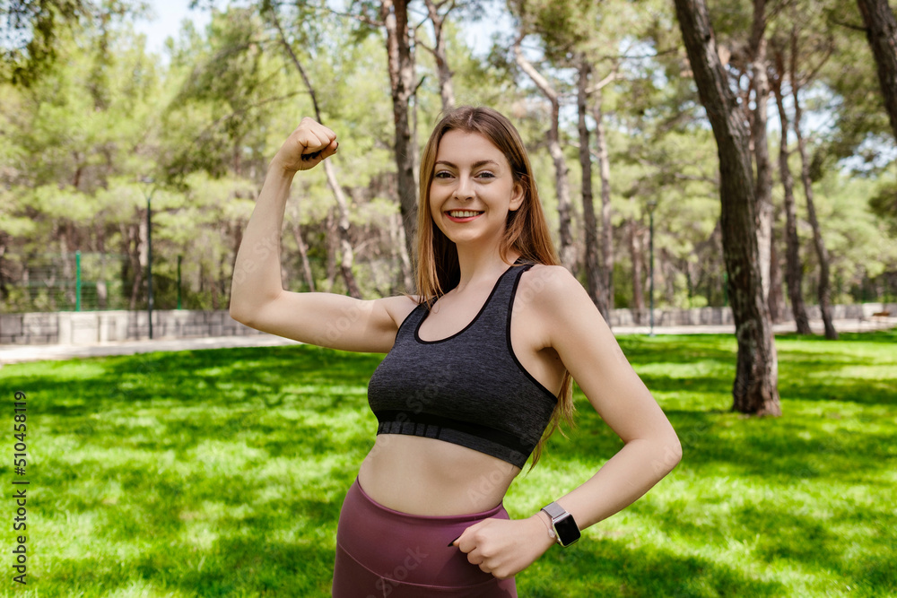 Brunette sportive woman wearing black sports bra standing on city park, outdoors showing her bicep and smiling. Healthy life and outdoor sport concepts.