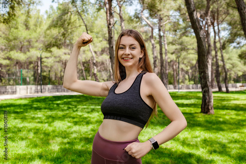Brunette sportive woman wearing black sports bra standing on city park, outdoors showing her bicep and smiling. Healthy life and outdoor sport concepts.