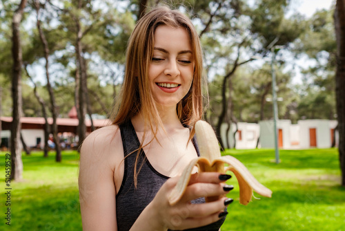 Cute brunette woman wearing sports bra standing on city park  outdoors eating healthy banana for strength and energy. Outdoor sports and healthy food and life concepts.