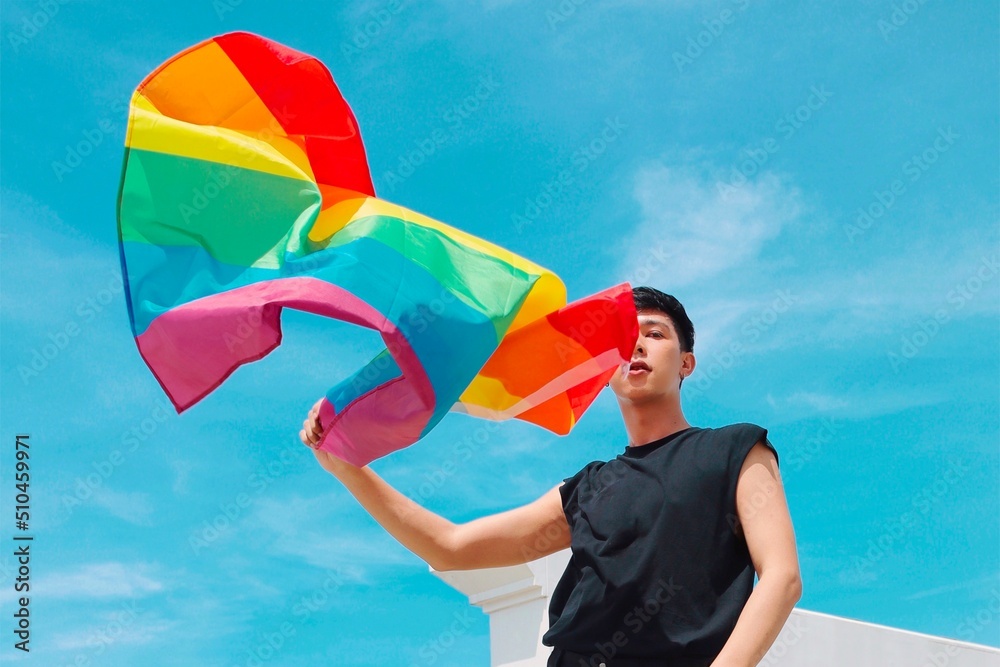Young man  standing over beautiful blue sky and waving lgbti+ flag.