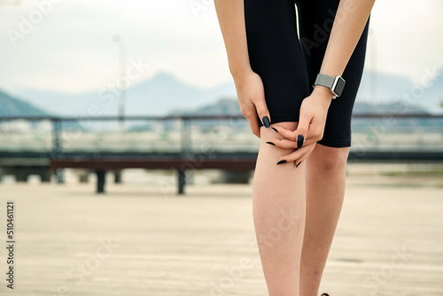 Young caucasian woman wearing black sports bra standing on city park, outdoors cramps in leg and feel pain, touching her knee ankle painful injury outside after exercise workout. Injuries concept.