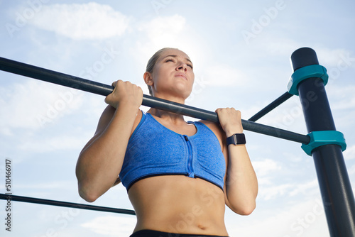 Below view of concentrated powerful young woman with fitness tracker hanging on horizontal bar while doing pull-up at workout area