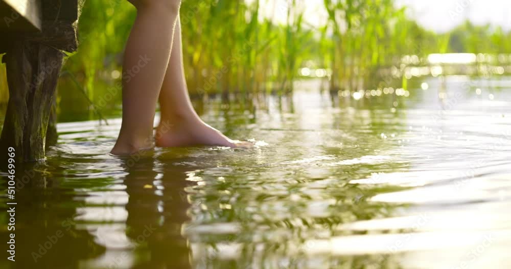 Closeup of children legs which are swinging, while touching water in a lake in a relaxed way. A child is sitting on the edge of a wooden pier in summer Closeup of children legs which are swinging, while touching water in a lake in a relaxed way. A child is sitting on the edge of a wooden pier in summer