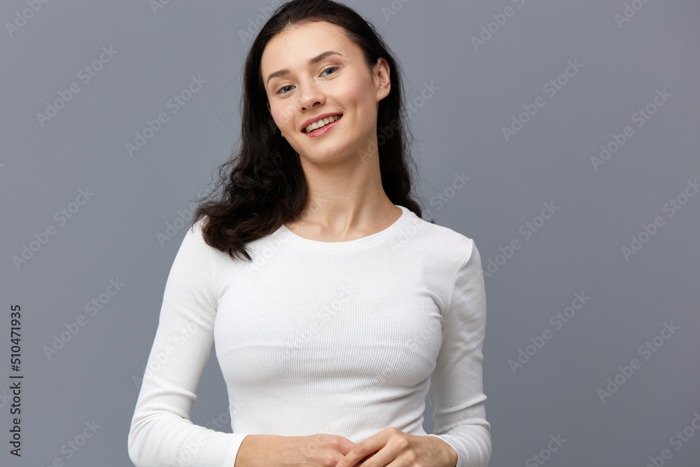 a sophisticated woman stands on a gray background in a white T-shirt, tilted her head to the side, dropping her hands down and joining them together a sophisticated woman stands on a gray background in a white T-shirt, tilted her head to the side, dropping her hands down and joining them together