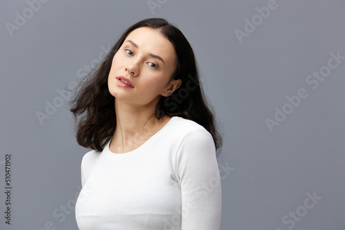 an attractive woman is standing on a gray background in a white T-shirt, with her hands behind her back, turning her head in different directions in a relaxed manner
