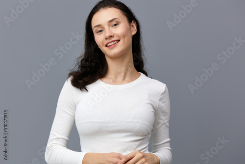 a sophisticated woman stands on a gray background in a white T-shirt, tilted her head to the side, dropping her hands down and joining them together