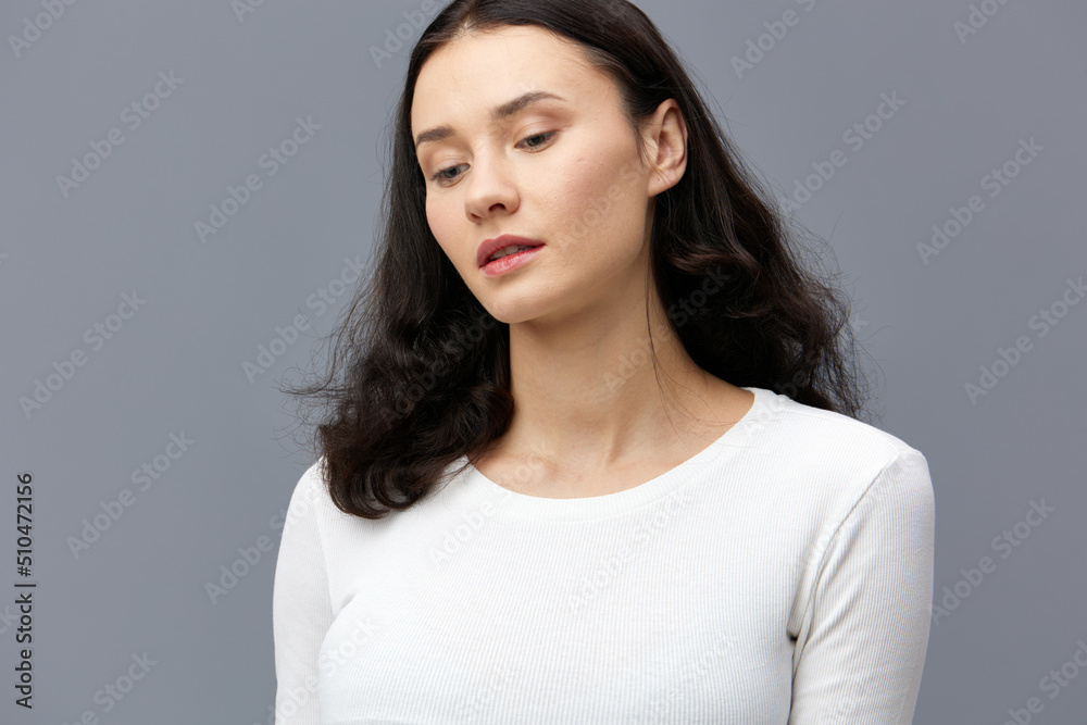 an attractive woman is standing on a gray background in a white T-shirt, with her hands behind her back, turning her head in different directions in a relaxed manner