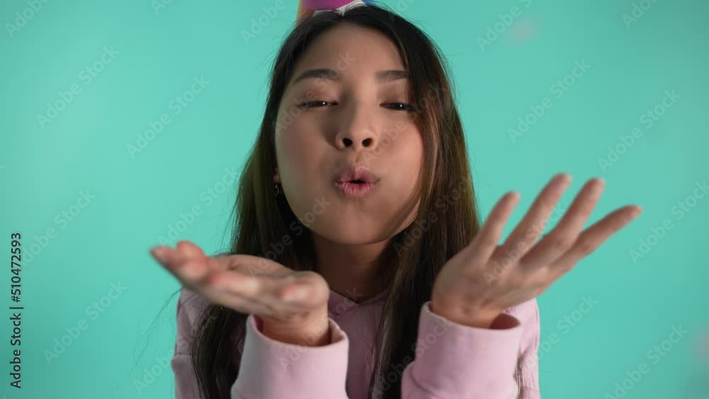Close-up of happy asian birthday girl wearing festive hat blowing colored sparkles isolated on blue background. Cheerful young woman smiling while looking at flying sequins during Birthday celebration