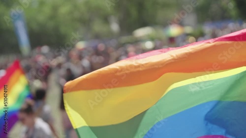 The LGBTQ rainbow flag waving in slow motion with people in a pride parade in the background