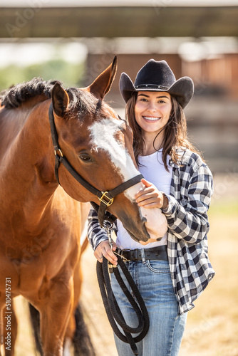 Good-looking young woman in cowboy hat strokes a horse on chops while smiling at the camera