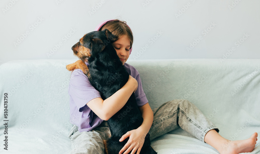 Happy teenage girl in lilac t-shirt sits on the sofa, listens to music has fun playing with the dog Happy teenage girl in lilac t-shirt sits on the sofa, listens to music has fun playing with the dog