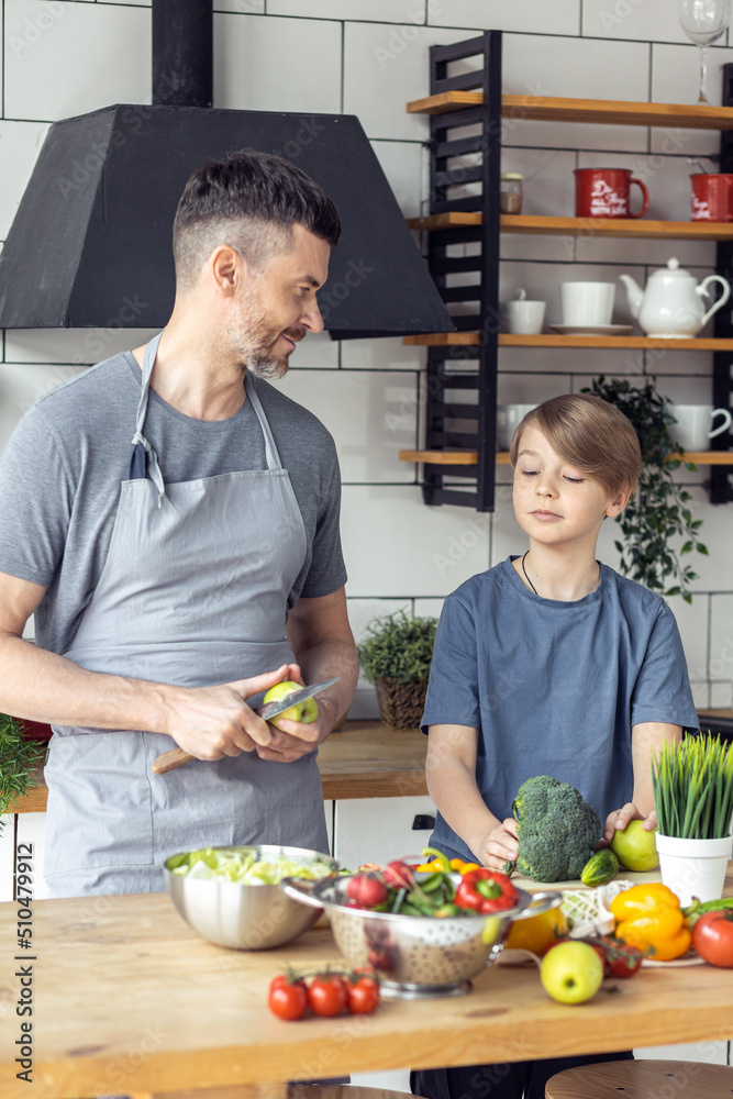 Handsome father and his teenager son spending quality time together. Men doing chores, cooking healthy vegetable salad, tasty food in the kitchen at home