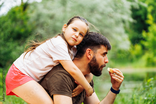 Side view of smiling family sitting on picnic in park forest around trees bushes. Little daughter with puffed cheeks sitting on fathers back. Middle-aged man eating food. Love, summer, togetherness. © Юля Бурмистрова