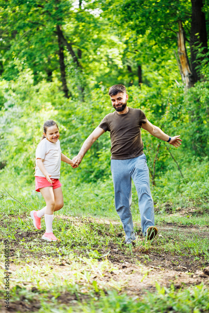 Portrait of happy family walking in park forest around green trees, having fun. Little cheerful daughter holding hand of middle-aged smiling bearded man father. Girl skipping. Love, summer activities. Portrait of happy family walking in park forest around green trees, having fun. Little cheerful daughter holding hand of middle-aged smiling bearded man father. Girl skipping. Love, summer activities.