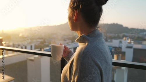 Woman starts her day with a cup of tea or coffee on the balcony at dawn, slow motion. City landscape in the background