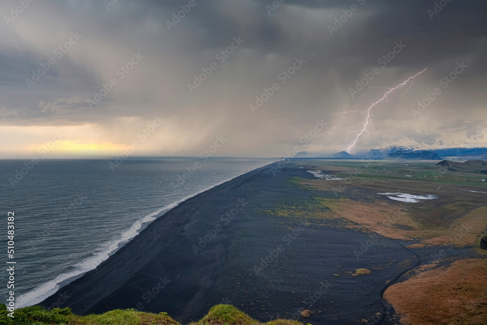 Scenic view of lightning weather over black sand beach. High angle view of sea waves rushing towards shore against dramatic sky. Beautiful landscape by mountains during sunset.