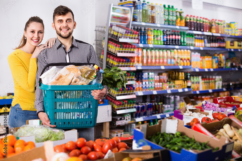 Portrait of smiling glad friendly couple with shopping basket filled food products in supermarket