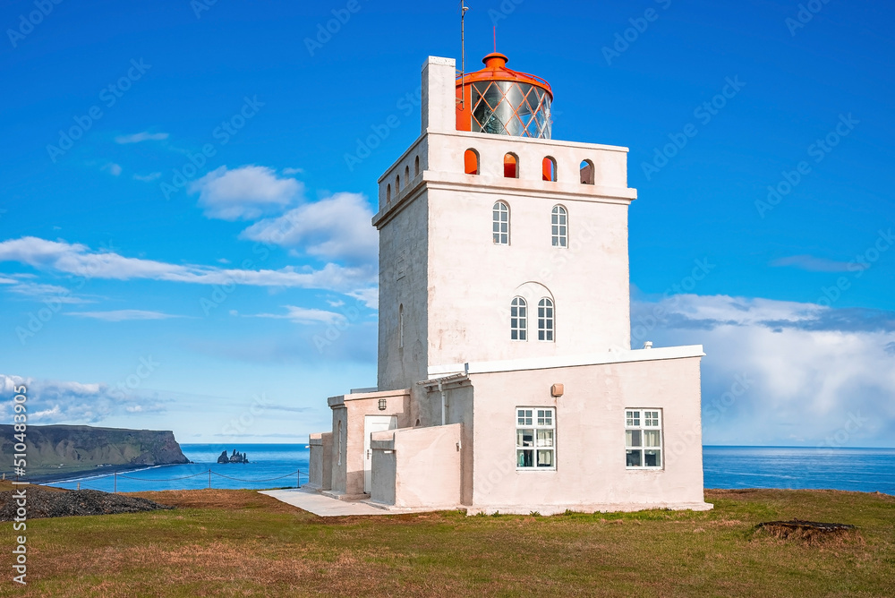 Beautiful Dyrholaey lighthouse on cliff during sunny day. Idyllic view of white concrete building at seashore in Dyrholaeyjarviti. Scenic view of Reynisfjara beach against blue sky.