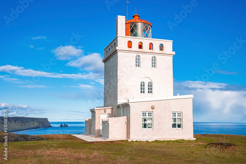 Beautiful Dyrholaey lighthouse on cliff during sunny day. Idyllic view of white concrete building at seashore in Dyrholaeyjarviti. Scenic view of Reynisfjara beach against blue sky.