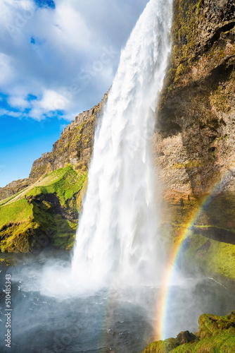 Beautiful Seljalandsfoss with rainbow flowing from mountain. Low angle view of majestic idyllic waterfall against cloudy sky. Scenic view of famous tourist attraction in valley.