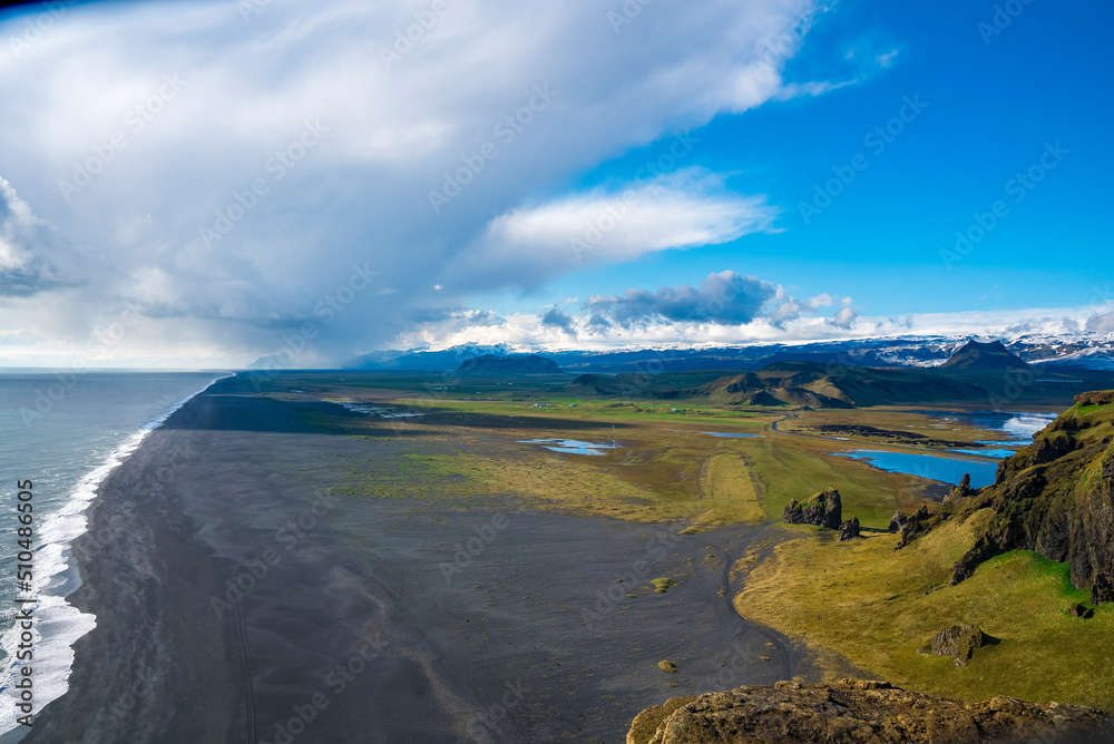 Idyllic view of black sand beach by mountains. High angle view of Dyrholaeyjarviti cliff. Beautiful picturesque scenery of volcanic landscape against blue cloudy sky . Idyllic view of black sand beach by mountains. High angle view of Dyrholaeyjarviti cliff. Beautiful picturesque scenery of volcanic landscape against blue cloudy sky .