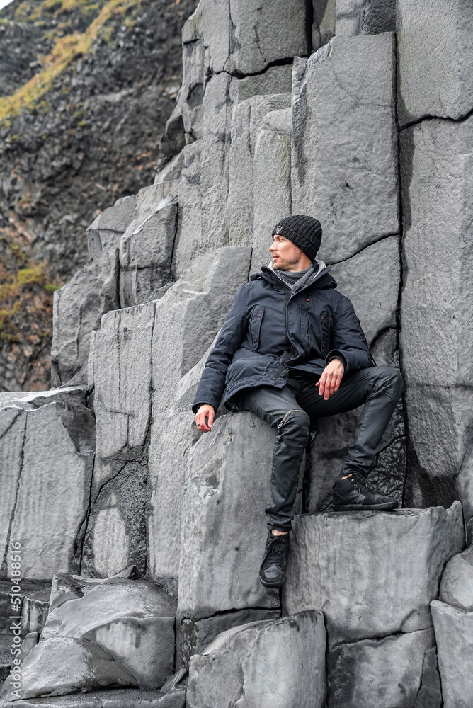 Thoughtful tourist sitting on beautiful basalt columns. Man looking away while relaxing on rocky formation. Scenic view of gray patterned stones on seashore at famous Reynisfjara Beach. Thoughtful tourist sitting on beautiful basalt columns. Man looking away while relaxing on rocky formation. Scenic view of gray patterned stones on seashore at famous Reynisfjara Beach.