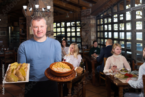 Hospitable owner of cosy country restaurant meeting guests with traditional meals in hands