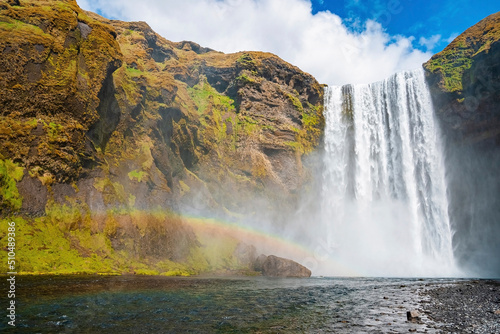 Low angle view of powerful cascades of Skogafoss waterfall. Beautiful river flowing amidst cliffs in valley. Idyllic natural scenery of volcanic landscape against blue sky.