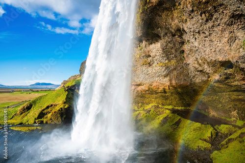 View of beautiful Seljalandsfoss with rainbow flowing from mountain. Majestic idyllic waterfall against cloudy sky. Scenic close-up view of famous attraction in valley.