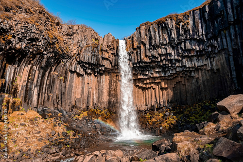 Beautiful Svartifoss waterfall amidst basalt rock columns formation. Stream flowing through rocks from cliffs against blue sky. Scenic view of famous tourist attraction in national park.