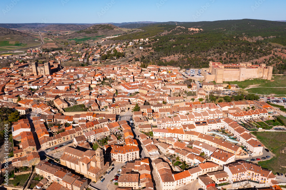 General aerial view of Siguenza city with brownish tiled roofs of residential houses, medieval cathedral and fortified castle surrounded by green hills on sunny spring day, Spain..