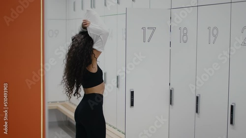 Young lady athlete taking off her t-shirt, getting ready for fitness workout, standing in front of locker at gym