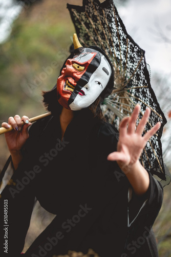 Portrait of sexy and young japanese woman with beautiful old traditional black kimono, traditional double mask (of 'hannya' and 'noh') and lace sun umbrella in the autumn forest  photo