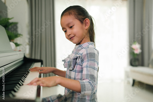 Cute asian 6 years old is practice playing piano at home and smiling with happiness moment, concept of learning, art, steam, musical, mental health, homeschool, skill, ability concept.