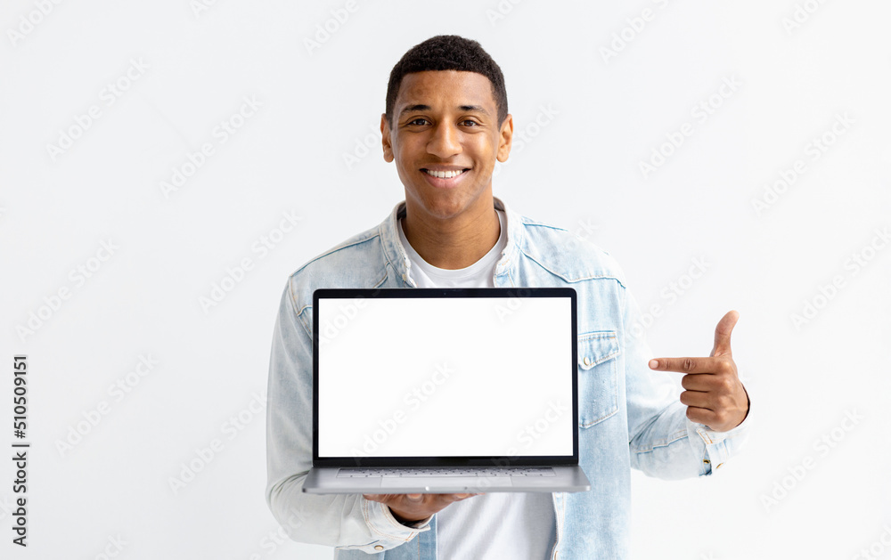 Portrait of young African American man with laptop on white background. Male points her finger at a blank laptop screen, looking at the camera and smiling Portrait of young African American man with laptop on white background. Male points her finger at a blank laptop screen, looking at the camera and smiling