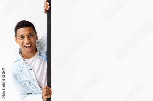 Young African American man peeking out from empty white board, looks at the camera and smiles. Copy space. Place for your advertising, mock-up
