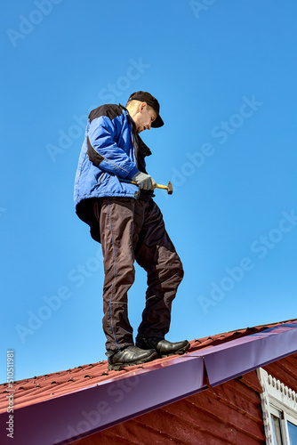 Roofer stands on roof with hammer in his hand against background of blue sky.