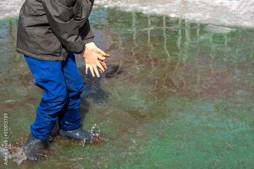 Joyfull Child in rubber boots kneading dirt with hands in muddy puddle. Environmental pollution. Child boy of 6-7 year old walking and playingin wellies in polluted water photo