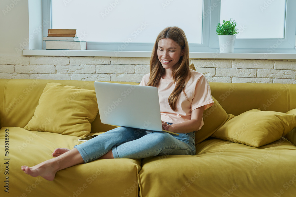Naklejka premium Beautiful young woman working on laptop and smiling while sitting on the couch at home