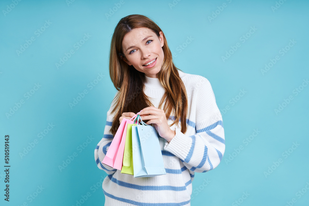 Beautiful young woman carrying little shopping bags and smiling while standing against blue background Beautiful young woman carrying little shopping bags and smiling while standing against blue background