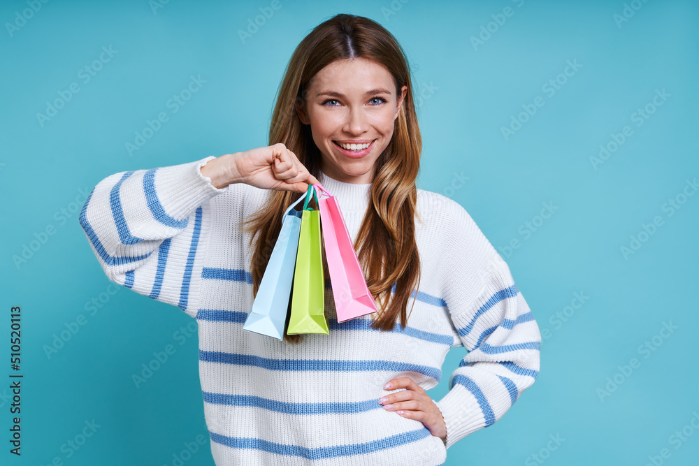 Beautiful young woman carrying little shopping bags and smiling while standing against blue background Beautiful young woman carrying little shopping bags and smiling while standing against blue background