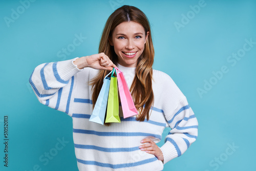 Beautiful young woman carrying little shopping bags and smiling while standing against blue background