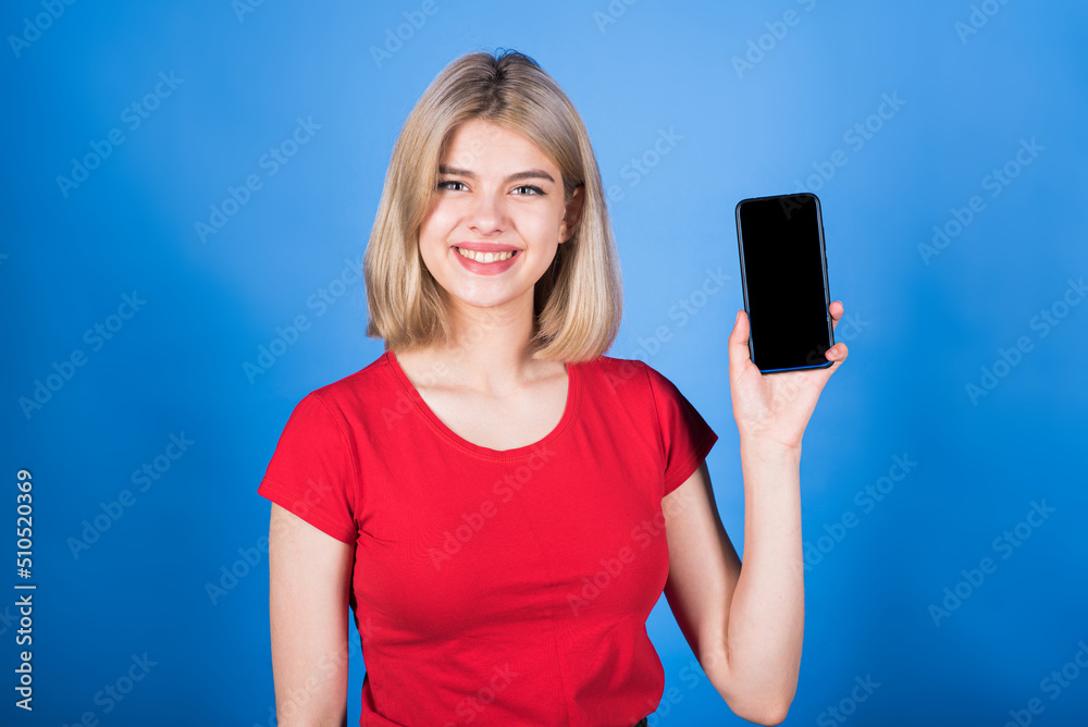 Young and attractive caucasian blonde girl in casual clothes showing the screen of a mobile phone with copy space for advertising isolated on a blue studio background. Young and attractive caucasian blonde girl in casual clothes showing the screen of a mobile phone with copy space for advertising isolated on a blue studio background.