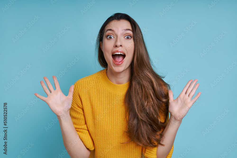 Surprised young woman gesturing and looking at camera while standing against blue background Surprised young woman gesturing and looking at camera while standing against blue background