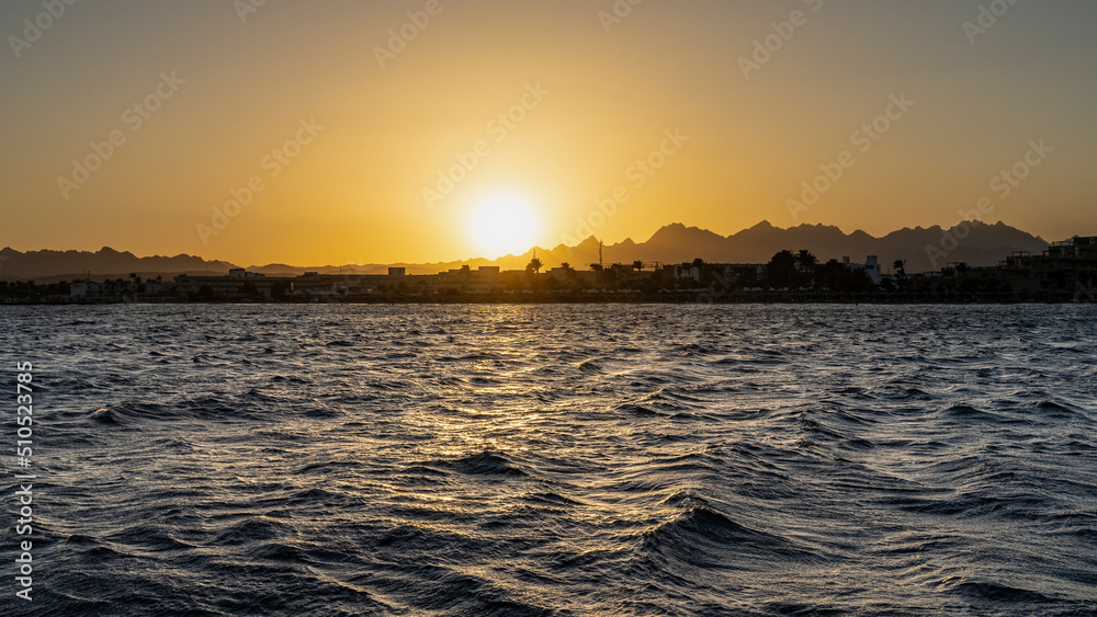 Sunset over the Red Sea. Egypt. Silhouettes of a mountain range against an orange sky. The sun is low. Waves on the water. Safaga.