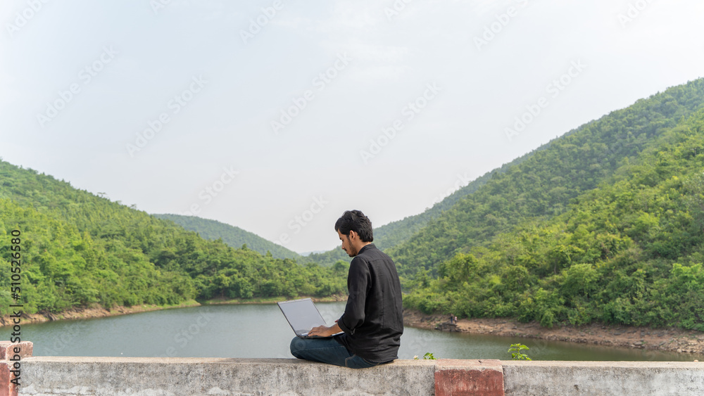 Fototapeta premium Young man working on a laptop and sitting in the nature, Workation and Digital Nomad concept.