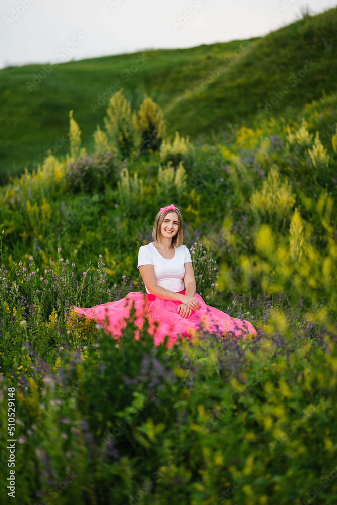 Princess in a summer pink dress sits on the green grass. The girl in a flower field in a country.